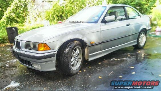 Silver BMW 3 Series coupe parked on wet ground, trees in background