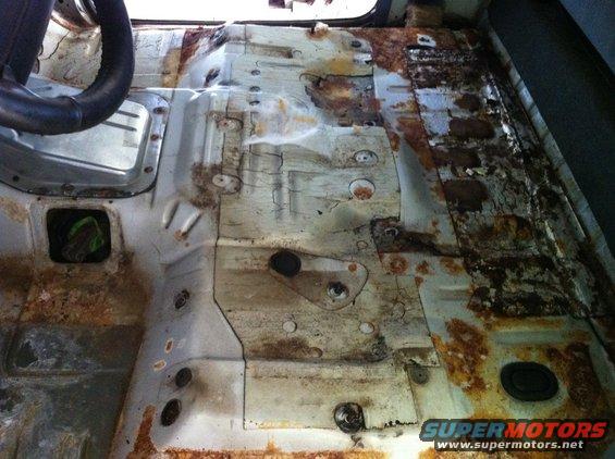 Vehicle interior floor with extensive rust and damage, showing a steering wheel and metal bracing