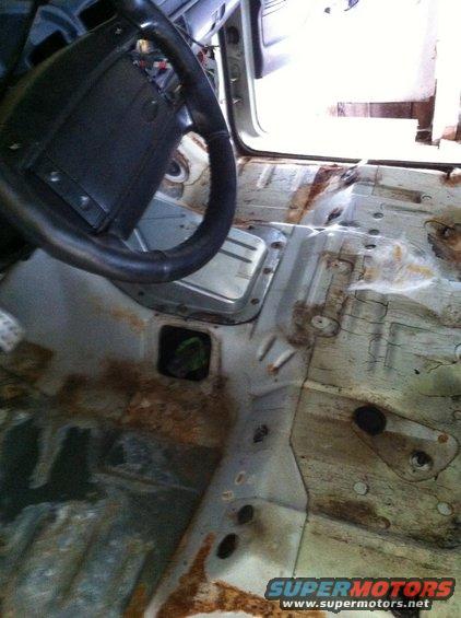 Interior floor of a vehicle showing significant rust and damage, with a steering wheel visible