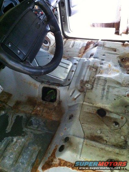 Interior of a vehicle with the floor pan heavily rusted and corroded, showing the steering wheel and dashboard area