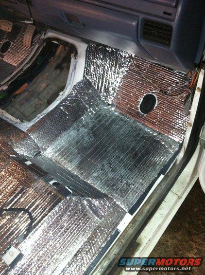 Vehicle interior floor with silver insulation installed, showing pedals and dashboard