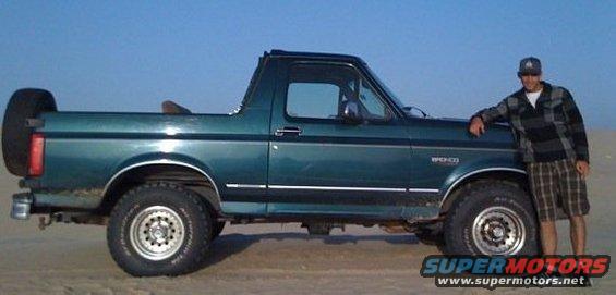 Green Ford Bronco with a spare tire on the back, parked on sand with a man standing next to it
