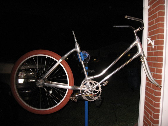 Chrome bicycle frame with a large red-orange tire on the rear wheel, leaning against a brick wall