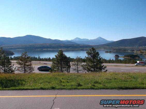 Car driving on highway next to a lake with mountains in the background. A truck is also visible on the highway