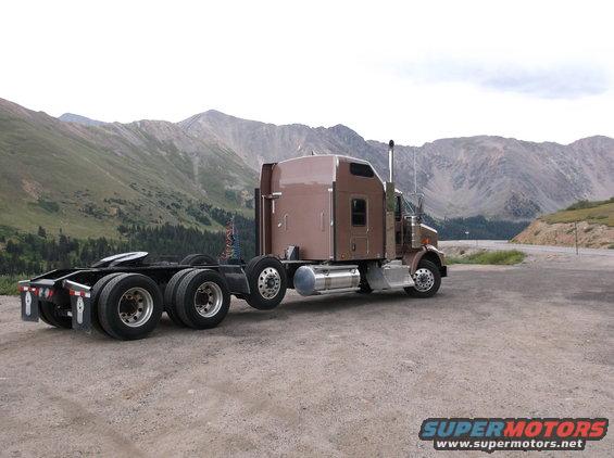 Brown semi-truck cab and chassis parked on a gravel road with mountains in the background