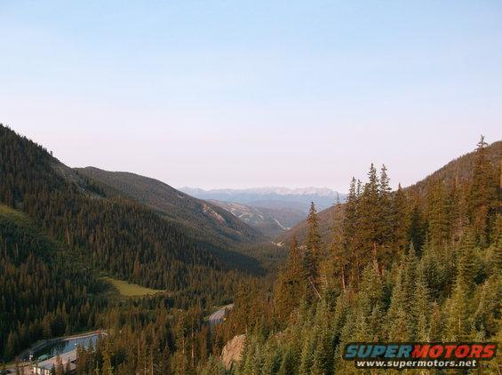 Mountain valley with dense evergreen trees, a winding road, and a body of water
