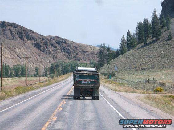 Rear view of a dump truck driving on a highway with mountains and trees in the background