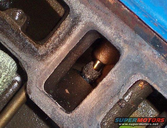 Close-up of an engine valve and its housing, showing wear and oil residue. A green and blue watermark reads 'SUPERMOTORS.NET'