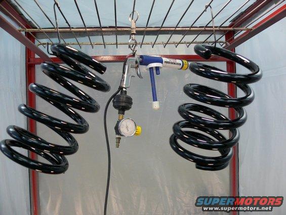 Two black coil springs hang from a rack, with a powder coating gun and pressure gauge nearby