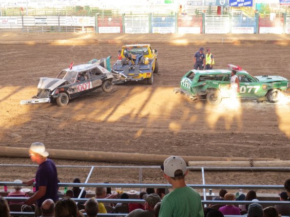 Demolition derby cars, one black with '007' and one green with '077', in a dirt arena with a tow truck