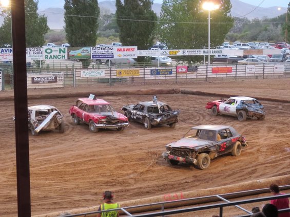 Several wrecked cars are on a dirt track during a demolition derby event. Spectators are visible in the foreground