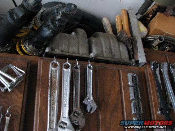 Engine intake manifolds and shock absorbers on a shelf above hanging wrenches and tools