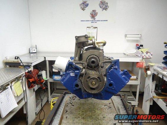 Blue engine block with timing gears and chain visible, suspended in a workshop