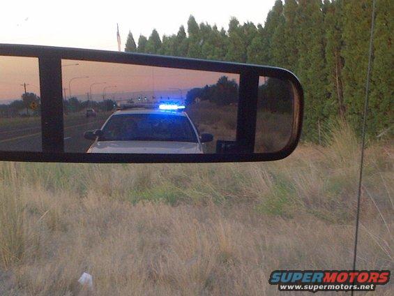 Police car with blue lights on reflected in a side mirror, driving on a road at dusk