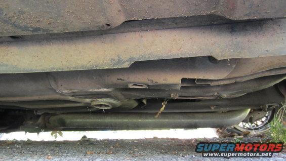 Underside view of a car, showing the exhaust system and frame components. A wheel is partially visible on the right
