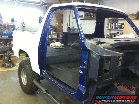 Truck body being worked on in a garage. White cab, blue doors, and bare metal chassis visible