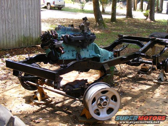 Jeep frame on jack stands with engine and one wheel attached. A white pickup truck is visible in the background