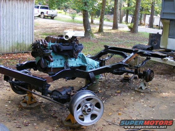 Vehicle frame and engine assembly on jack stands outdoors. A white pickup truck is visible in the background