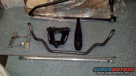 Various black and silver automotive suspension components laid out on a speckled floor. Includes sway bars and adjustable linkages