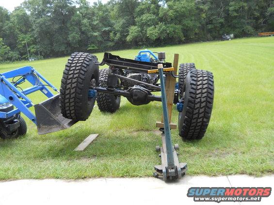 Vehicle chassis with large off-road tires, supported by a jack and a blue tractor loader
