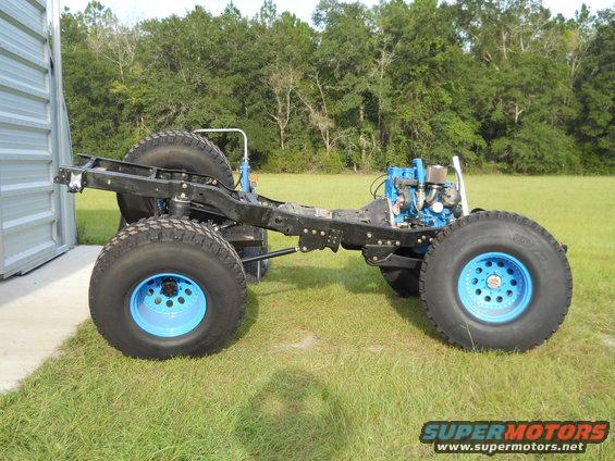Off-road vehicle chassis with blue engine, large tires, and bright blue rims. Next to a metal building