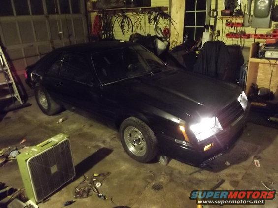 Black 1981 Ford Mustang coupe parked in a garage with the headlights on