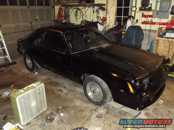 Black coupe car in a garage, front 3/4 view, with aftermarket wheels and a modified grille