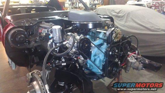 Car engine bay with a blue engine block, black hood scoop, and red fender. Another car is covered in a tarp in the background