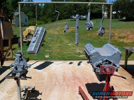 Engine block on stand, fuel tank hanging, and other car parts painted gray outdoors