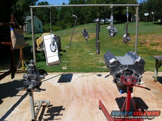 V8 engine on stand, transmission, and other car parts hanging from a rack outside
