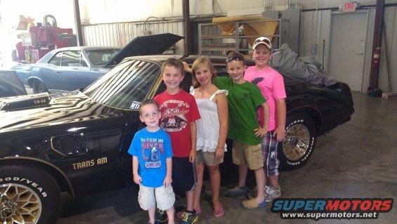 Kids pose in front of a black Pontiac Trans Am. Another car is in the background