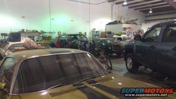 Gold car with black stripes in foreground, garage with several cars and a truck