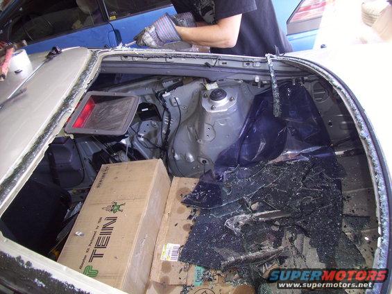 Car interior with shattered rear window, boxes, and debris. A person in gloves works on the car