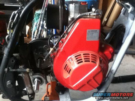 Close-up of a red engine component with vents and a silver label. Some metal tubing and black frame parts are visible