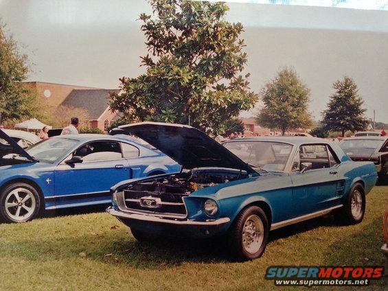 Blue 1967 Ford Mustang coupe with hood open, parked next to a newer blue Mustang. Cars are on grass at an outdoor event