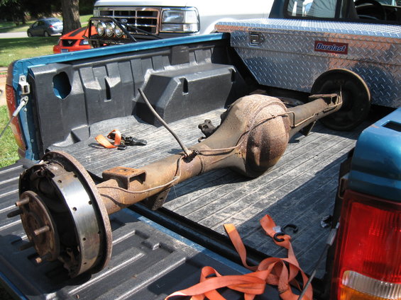 Rear axle assembly with brake drums and straps in the bed of a teal pickup truck. A white Ford truck is behind it