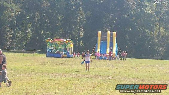 Two inflatable bounce houses and a large inflatable slide are set up on a grassy field with trees in the background. Several people, including children, are visible near the inflatables