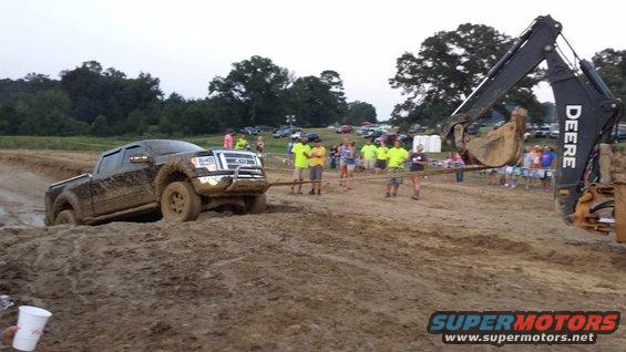 Mud-covered 2009 Ford F-150 stuck in a mud pit, with a Deere excavator pulling it. Spectators watch