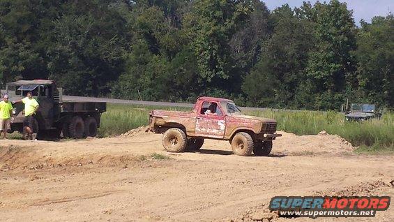 Muddy red pickup truck with a tow rope attached, being pulled by a military flatbed truck. Another vehicle is visible in the background