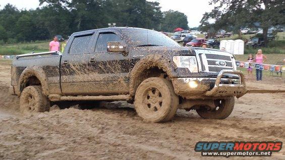 Muddy pickup truck driving through a dirt track with people and other vehicles in the background