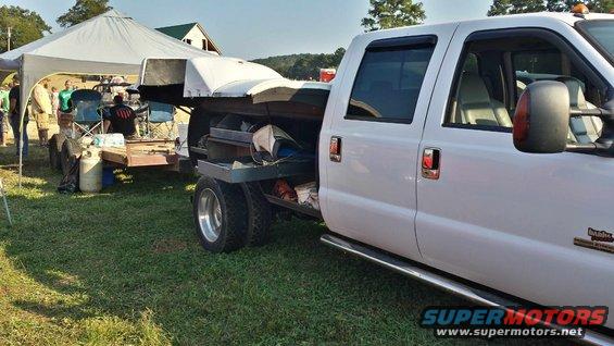 White pickup truck with a flatbed and dual rear wheels, towing a trailer with a tent and people in the background