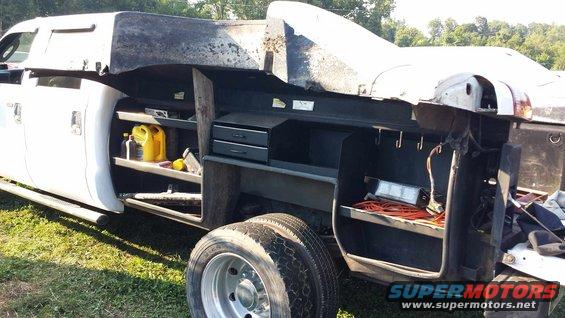 Service truck bed with shelves holding yellow jug, tools, and orange cords. Dual rear wheels visible