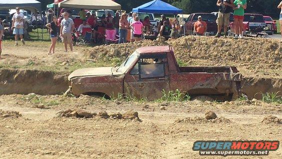 Red pickup truck stuck in a mud pit, with spectators watching from higher ground