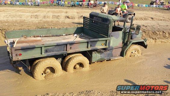 Camouflaged military flatbed truck stuck in mud pit with people watching