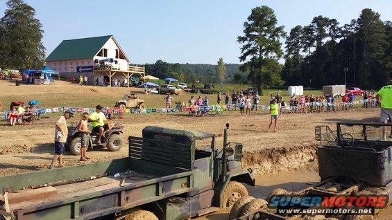 Mud bogging event with military trucks, ATVs, and spectators gathered around a muddy track. A barn is visible in the background