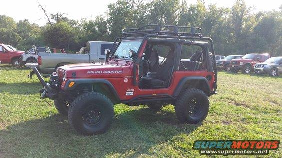 red Jeep Wrangler with roof rack and off-road tires, parked in a grassy field with other trucks and SUVs in background