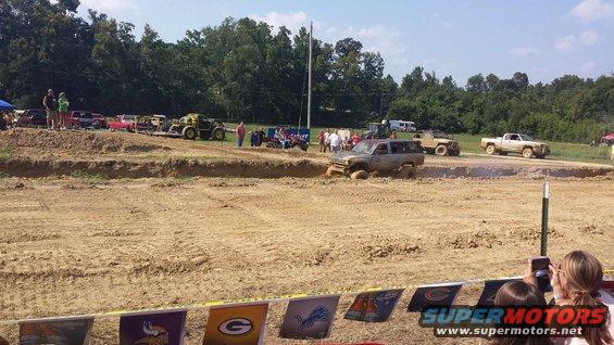 Several off-road vehicles are navigating a mud pit during an event. One SUV is stuck in the mud, while a pickup truck drives past