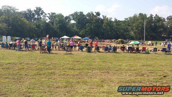 Outdoor event with people sitting in chairs and several vehicles in the background