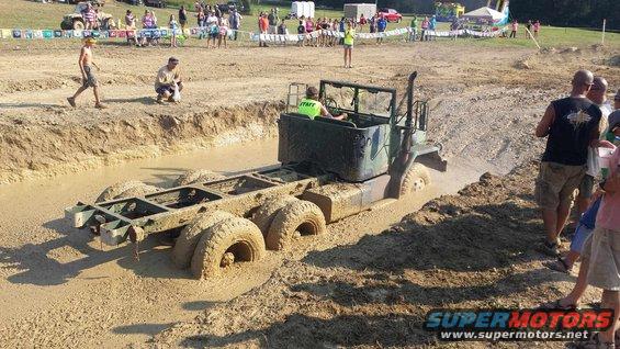 Military truck chassis with multiple axles stuck deep in mud. Driver wearing a green 'STAFF' shirt is visible in the cab. Spectators watch from the bank