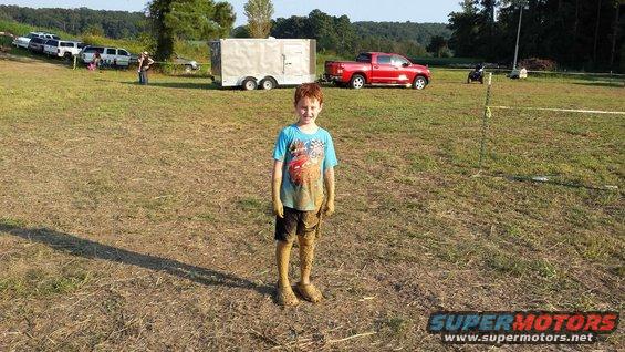 Mud-covered boy standing in a grassy field with a red pickup truck and trailer in the background